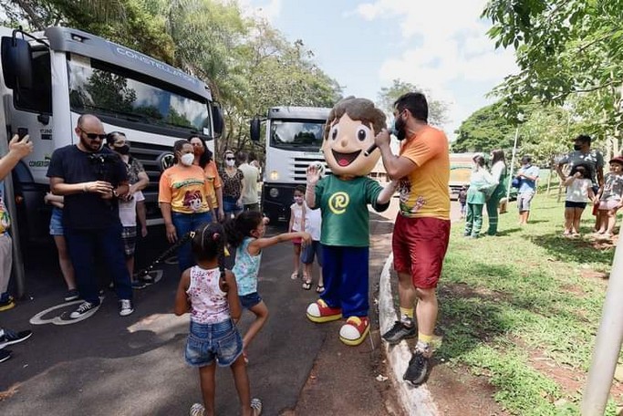 Mascote Recicladinho cumprimentando crianças ao lado de caminhões de coleta seletiva
